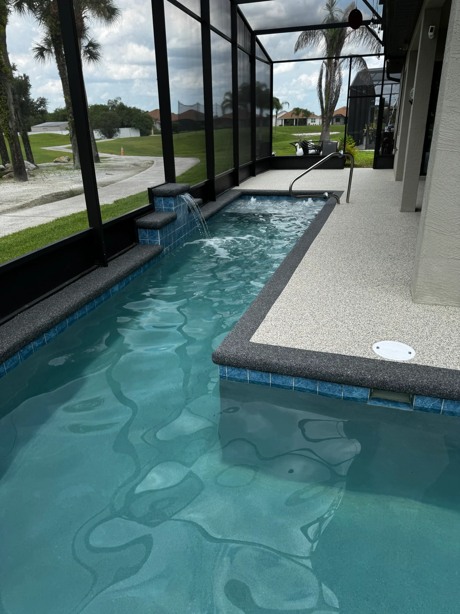 Speckled gray concrete pool deck with blue tile border and waterfall feature in screened enclosure